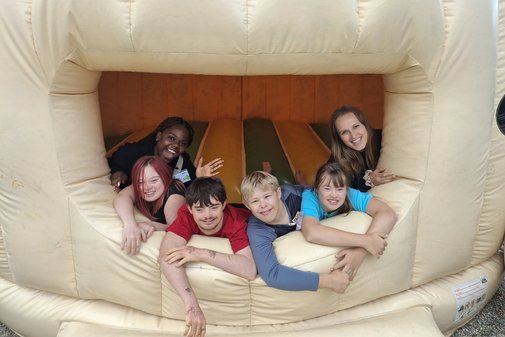 Zwei Freizeitbegleiterinnen und vier Kinder, zwei Mädchen und zwei Jungen, schauen aus dem Eingang zu einer Hüpfburg. Sie lachen.