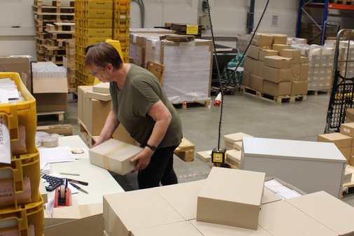 Eine Mitarbeiterin von Dimetria hebt ein Paket von ihrem Arbeitsplatz. Um sie herum stehen Paletten mit Paketen und gelbe Postkisten.