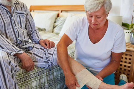 Eine ältere Frau legt einer Person, die im Pyjama auf dem Rand eines Bettes sitzt, einen Verband an der linken Wade an. Die Frau kniet dazu vor der Person, der Kopf der anderen Person ist nicht zu erkennen.