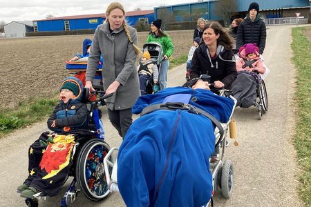 Die Szene zeigt eine Straße und daneben einen Acker, im Hintergrund ein blaues Industriegebäude. Mehrere Frauen schieben die sieben pflegebedürftigen Kinder und Jugendlichen in ihren Kinderwägen sowie einen Jugendlichen im Pflegebett.