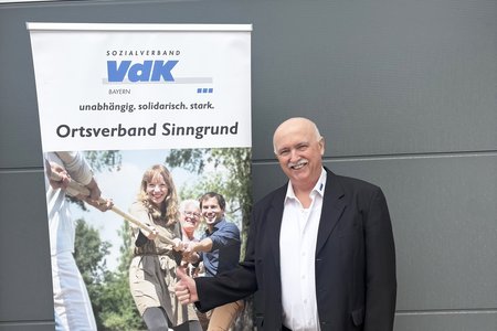 Das Foto zeigt den Ehrenamtlichen Anton Breitenbach vor einem Roll-up des VdK-Ortsverbands Sinngrund. Er lächelt.