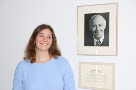 Eine junge Frau steht an einer weißen Wand, an der ein Schwarz-Weiß-Foto eines älteren Mannes hängt. Sie lächelt. Das Bild zeigt Karoline Weishäupl neben dem Bild ihres Großvaters Karl Weishäupl, der den VdK zusammen mit anderen 1946 ins Leben gerufen hat.