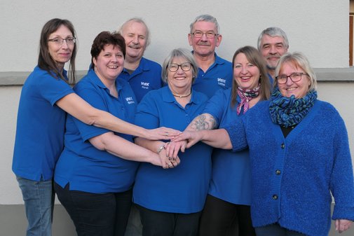 Acht VdK-Ehrenamtliche stehen eng nebeneinander und legen ihre Hände übereinander. Sie tragen blaue VdK-T-Shirts und lachen in die Kamera.