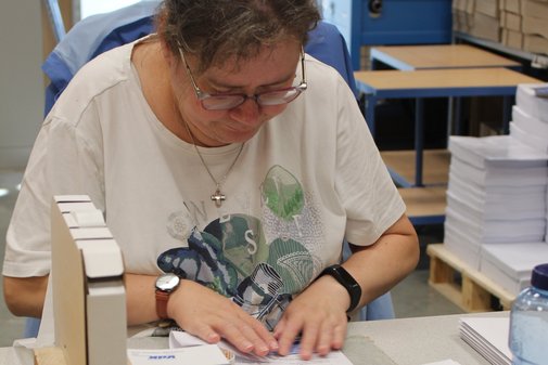 Eine Mitarbeiterin von Dimetria sitzt an ihrem Arbeitsplatz und faltet einen Brief. Vor ihr liegen VdK-Broschüren und Briefumschläge.