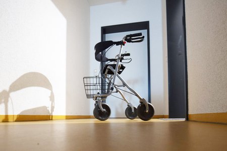 Das Bild zeigt einen Rollator, der in einem Hausgang steht. Krankenkassen finanzieren nur günstige Rollatoren, die schwer und unhandlich sind. Wer damit nicht zurechtkommt, muss sich auf eigene Kosten einen Rollator beschaffen. Viele Pflegebedürftige brauchen das Landespflegegeld, um Notwen¬diges anzuschaffen, etwa einen leichten Rollator.
