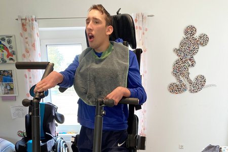 Louis Baumann, ein junger Mann mit Behinderung, steht in einem Stepper. Er legt die Hände auf die Handgriffe des Geräts, und die Füße sind mit Gurten fixiert. Im Hintergrund ist ein Rollstuhl zu sehen.