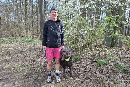 Ultraläuferin Sandra Spörl trägt sportliches Outfit und Käppi. Sie hält ihren altdeutschen Hütehund Janosch an der Leine. Beide stehen auf einem Wanderweg im Meilwald an einem sonnigen Tag. Links im Hintergrund viele Bäume, rechts ein blühender Busch.