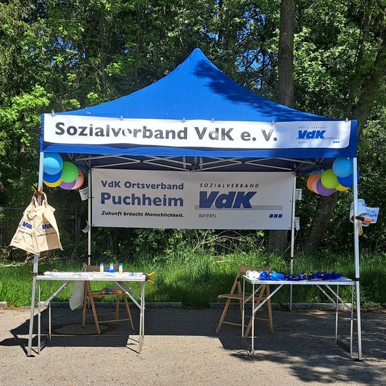 Der VdK Infostand wartet in Puchheim auf neue Besetzer Ein blauer Pavillon durch weiße Banner als Infostand des VdK Puchheim kenntlich gemacht. Er ist mit Luftballons und Fähnchen geschmückt. Darunter sind 2 Tische mit Info- und Werbematerial, sowie zwei leere Klappstühle aufgestellt.