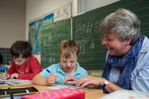 Eine Frau (rechts im Bild) sitzt in einem Klassenzimmer neben einem Jungen mit Down-Syndrom und hilft ihm bei einer Aufgabe.